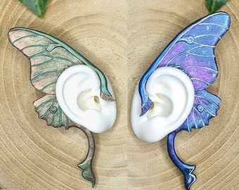 May include: A pair of ear cuffs shaped like moth wings. The left cuff features metallic green and bronze tones with detailed veining. The right cuff displays a vibrant blue and purple gradient with white star accents. Both cuffs wrap around white ear models.