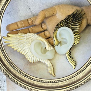Könnte beinhalten: Ein Paar Ohrklemmen mit Flügeldesign. Eine Manschette ist gold und weiß, die andere gold und schwarz. Sie sind vor einer Holzhand und einem goldgerahmten Hintergrund platziert. Die Ohrklemmen sind für das Ohr konzipiert.