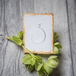 May include: A white card with gold edges and the number "3" written in black calligraphy. The card is lying on a wooden surface with green leaves.