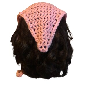 Op de afbeelding: Een roze gehaakte bandana met een driehoekige vorm. De bandana is om het hoofd van een persoon gebonden.