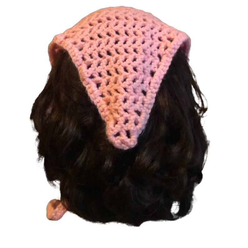 Op de afbeelding: Een roze gehaakte bandana met een driehoekige vorm. De bandana is om het hoofd van een persoon gebonden.