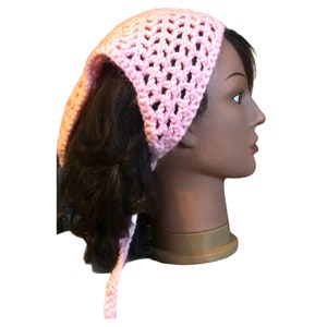 Op de afbeelding: Een roze gehaakt hoofdband met een lange strik. De hoofdband is gemaakt met een eenvoudig steekpatroon en heeft een losse, luchtige uitstraling.