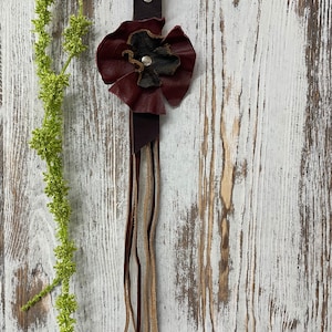 Puede incluir: Un llavero de cuero burdeos con un adorno floral y flecos de cuero colgantes. La flor tiene un centro verde oscuro y está sujeta a una correa de cuero marrón oscuro con un cierre de metal plateado. El fondo es una superficie de madera blanqueada.