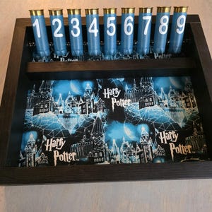 May include: A dark wooden display case featuring nine blue shotgun shells with white numbers 1-9. The bottom of the case has a Harry Potter themed design with a castle and the words "Harry Potter".