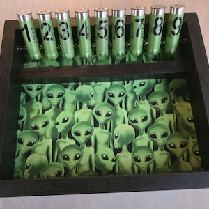 May include: A wooden box with a green alien-themed design. The box features a collection of green alien figures and nine green shotgun shells labeled with numbers 1 through 9. The shells are arranged in a row above the alien figures.