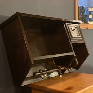 Może przedstawiać: Drewniana p&oacute;łka ścienna w ciemnym kolorze brązu z małym sejfem po prawej stronie. P&oacute;łka ma mały otwarty przedział po lewej stronie i metalową rurę z dziurką od klucza na dole. P&oacute;łka jest zamontowana na ścianie.