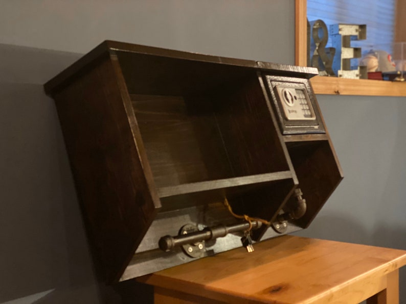 Może przedstawiać: Drewniana p&oacute;łka ścienna w ciemnym kolorze brązu z małym sejfem po prawej stronie. P&oacute;łka ma mały otwarty przedział po lewej stronie i metalową rurę z dziurką od klucza na dole. P&oacute;łka jest zamontowana na ścianie.