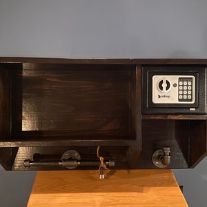 Może przedstawiać: Drewniana p&oacute;łka ścienna w ciemnym kolorze brązu z małym sejfem, dwoma metalowymi haczykami i metalową rurą do zawieszania przedmiot&oacute;w. Sejf ma cyfrowy panel sterowania i napis "ezSafe".