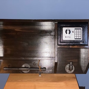 Może przedstawiać: Drewniana p&oacute;łka ścienna w ciemnym kolorze brązu z małym czarnym sejfem z cyfrową klawiaturą. Sejf ma nadrukowane słowo "Roadtop". P&oacute;łka ma metalową rurkę i dwa metalowe haczyki.