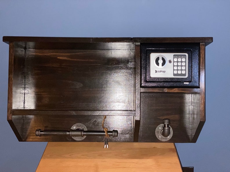 Może przedstawiać: Drewniana p&oacute;łka ścienna w ciemnym kolorze brązu z małym czarnym sejfem z cyfrową klawiaturą. Sejf ma nadrukowane słowo "Roadtop". P&oacute;łka ma metalową rurkę i dwa metalowe haczyki.