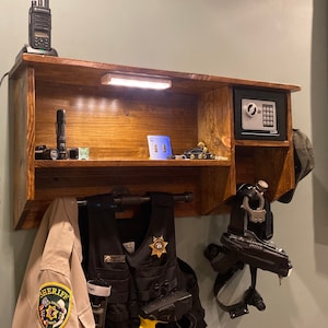 Może przedstawiać: Drewniana p&oacute;łka ścienna z sejfem, światłem i r&oacute;żnymi przedmiotami, w tym radiem, latarką i kamizelką. P&oacute;łka ma metalowy pręt do wieszania przedmiot&oacute;w, a na pręcie wisi kamizelka. Kamizelka ma naszywkę z napisem "Sheriff".