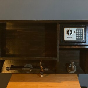 Może przedstawiać: Drewniana p&oacute;łka ścienna w ciemnym kolorze brązu z małym sejfem, drążkiem na ręczniki i dwoma haczykami. Sejf ma cyfrowy panel sterowania i napis "Sentry" na nim.