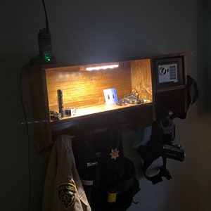 Może przedstawiać: Drewniana p&oacute;łka z lampką nad nią. Na p&oacute;łce znajdują się r&oacute;żne przedmioty, w tym latarka, radio i odznaka. Pod p&oacute;łką wiszą r&oacute;wnież ubrania na haczykach.