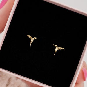 Könnte beinhalten: Ein Paar goldene Kolibri-Ohrringe in einer rosa Box. Die Ohrringe sind zart und haben ein detailliertes Kolibri-Design.