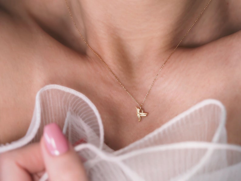 Pode incluir: Um delicado colar de ouro com um pingente de beija-flor. O colar &eacute; usado por uma pessoa com uma blusa branca e transparente.