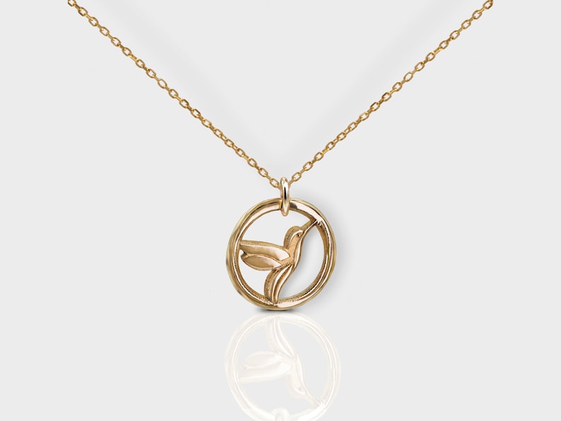 Pode incluir: Um colar de ouro com um pingente circular que apresenta um design detalhado de beija-flor.