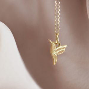 Pode incluir: Um colar com um pingente de beija-flor de ouro em uma corrente delicada. O pingente &eacute; uma representa&ccedil;&atilde;o estilizada e detalhada de um beija-flor em voo.