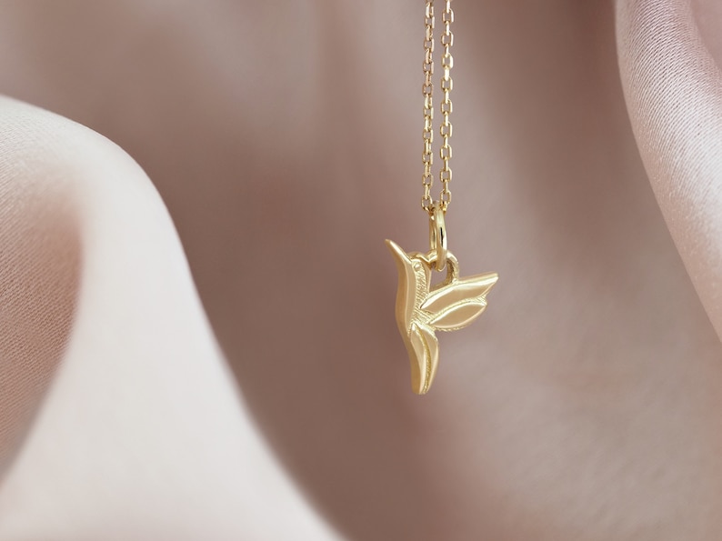 Pode incluir: Um colar com um pingente de beija-flor de ouro em uma corrente delicada. O pingente &eacute; uma representa&ccedil;&atilde;o estilizada e detalhada de um beija-flor em voo.