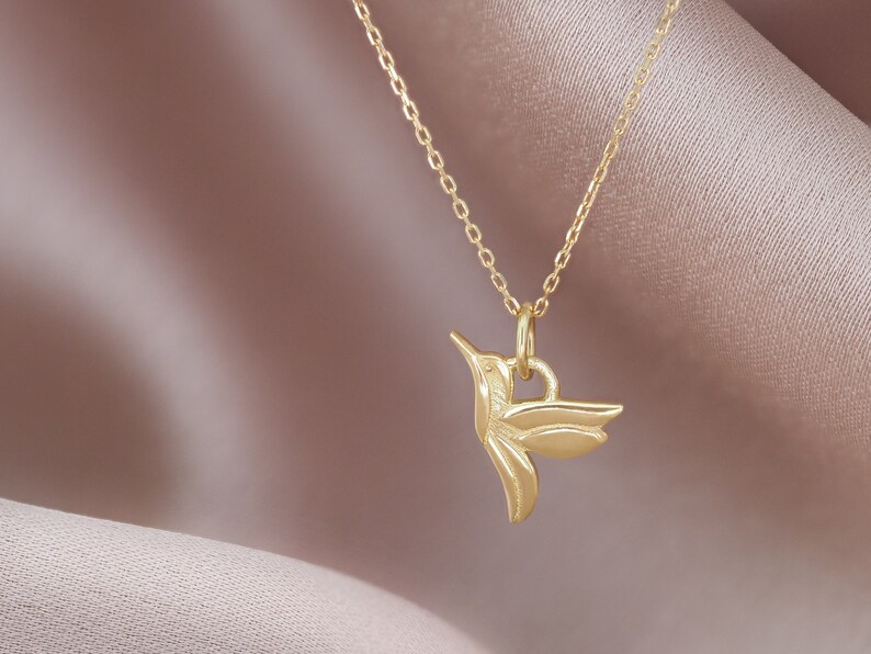 Pode incluir: Um colar de corrente de ouro com um delicado pingente de beija-flor. O beija-flor &eacute; detalhado com asas e um bico.