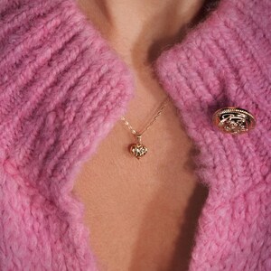 May include: A close-up of a pink knitted cardigan with a gold heart pendant necklace and a gold button. The cardigan has a cable knit pattern.