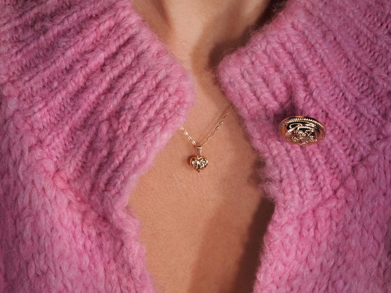 May include: A close-up of a pink knitted cardigan with a gold heart pendant necklace and a gold button. The cardigan has a cable knit pattern.