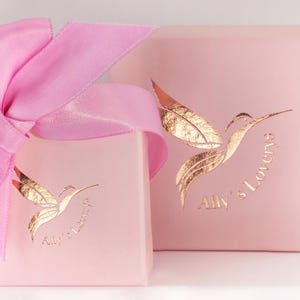 May include: Two pink gift boxes with a gold hummingbird logo and the text "Ally's Lovery's". The boxes are tied with a pink ribbon.