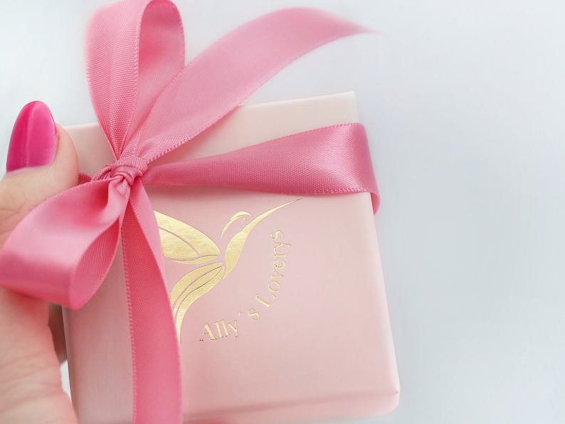 May include: A pink gift box with a pink satin ribbon tied in a bow. The box has a gold butterfly logo with the text "Ally's Lovery's".