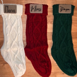 May include: Three knitted Christmas stockings with personalised leather name tags. The stockings are white, red, and green. The name tags read "Nana", "Mimi", and "Papa".