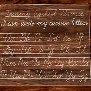 May include: A clear plastic dry-erase board with cursive letters and lines for practicing handwriting. The board has the text "Tommy Ezekiel Arinie I can write my cursive letters" at the top and the alphabet in cursive letters on the rest of the board.