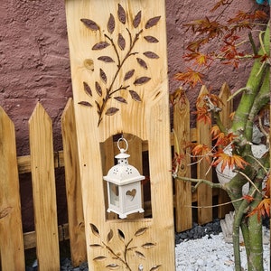 Könnte beinhalten: Ein hölzernes Willkommensschild mit geschnitzten Eichhörnchen und einer Laterne, die oben hängt. Das Schild ist auf einem Holzpfosten mit einem geschnitzten Blattdesign montiert. Eine Topfpflanze mit rosa und weißen Blüten steht auf einer Holzplattform unter dem Schild.