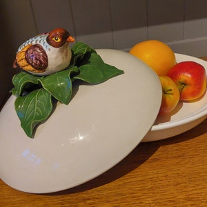 May include: A white ceramic serving dish with a lid, featuring a hand-painted bird figurine with green leaves. The dish is filled with red and orange fruit.