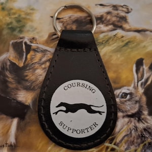 May include: Black leather keychain with a silver key ring. The keychain features a white circular emblem with the words "Coursing Supporter" and a black silhouette of a running dog. The background shows a painting of hares.