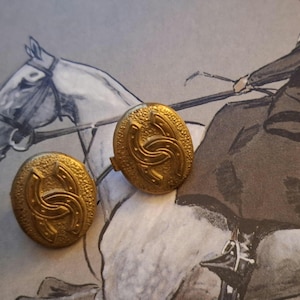 May include: A pair of antique gold buttons with a textured finish. Each button displays a design of two interlocking horseshoes. The buttons are circular and have a vintage appearance, possibly from a coat or jacket.