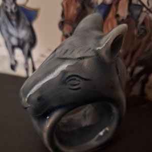 Puede incluir: Un anillo de servilleta de cerámica con forma de cabeza de caballo. El anillo es de color azul claro con detalles blancos y un diseño detallado.