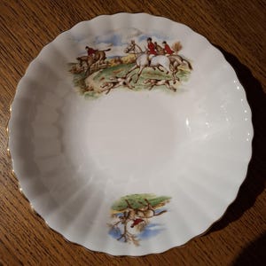 Puede incluir: Un tazón de cerámica blanca con un borde dorado y un diseño de escena de caza. La escena presenta jinetes a caballo con perros que persiguen a un zorro.