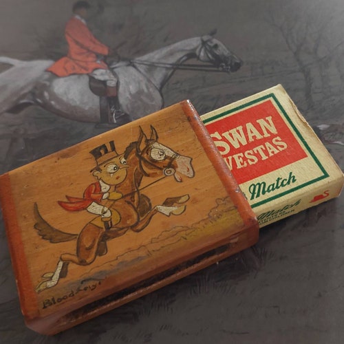 An art deco varnished wooden match box sleeve holds a standard Swan 2024 Vesta match box. Comical huntsman and a horse handpainted. 1920's style