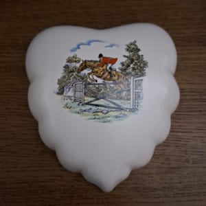 May include: Cream-colored, heart-shaped decorative plate with a detailed equestrian scene. The image depicts a rider in a red coat jumping a fence on a brown horse, set against a backdrop of trees and a blue sky.