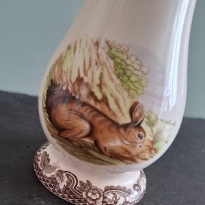 May include: A white ceramic vase with a brown rabbit illustration and green foliage. The vase has a brown floral pattern around the base.