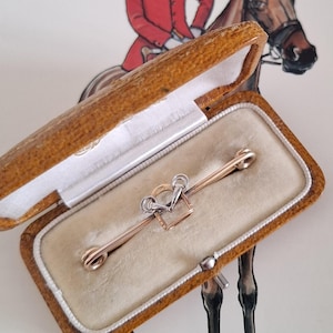 May include: A vintage gold and silver bar pin with a horse bit design, presented in a tan leatherette box with a cream-colored interior. The background features a drawing of a rider on a horse.