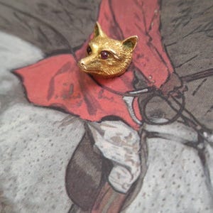 Puede incluir: Un broche con forma de cabeza de zorro dorada con ojos de piedras preciosas rojas. El broche de zorro es un diseño tridimensional detallado. El broche está sobre un fondo con tonos rojos y grises.