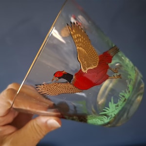 May include: A clear glass tumbler featuring a colourful illustration of a pheasant in flight. The bird has a red body, a black neck, and brown wings. Green grass is painted along the bottom edge of the glass, which has a thin gold rim at the top.