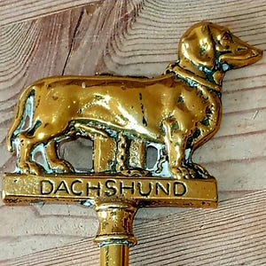 May include: A gold-toned metal decorative topper featuring a standing dachshund dog figure mounted on a horizontal bar with the word DACHSHUND embossed in capital letters. The metallic surface shows a polished finish with some dark patina.