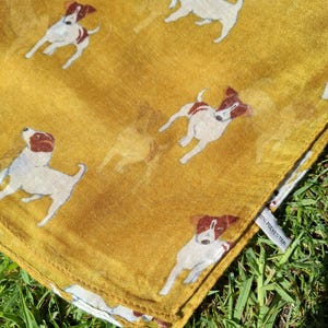 Puede incluir: Bufanda de color amarillo mostaza con un estampado repetido de perros blancos y marrones. La bufanda está hecha de un material ligero y tiene una etiqueta que dice "100% Poliéster". La bufanda está doblada y sobre hierba verde.