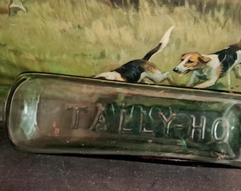 Op de afbeelding: Een doorzichtige, rechthoekige glazen fles met de woorden TALLY-HO in reliëf in hoofdletters op de voorzijde. De fles heeft een smalle hals en is horizontaal geplaatst voor een geschilderde scène met twee jachthonden.