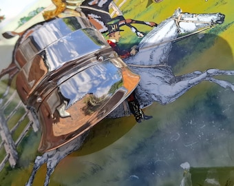 Op de afbeelding: Een gepolijste zilverkleurige metalen julepbeker met een glad, reflecterend oppervlak en een uitlopende rand. De beker is geplaatst over een kleurrijke illustratie van een paardenrace met een ruiter in een hoge hoed op een wit paard in een groen veld.