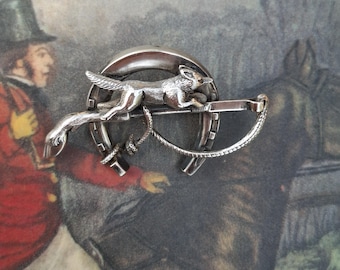 May include: A silver-toned metal brooch featuring a fox in a running pose positioned across a horseshoe. The design includes a thin, curved metal element resembling a riding crop or whip that extends across the center of the horseshoe shape.