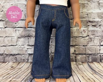 May include: Dark blue denim jeans for a doll, featuring a high-waisted design with visible orange contrast stitching, a faux fly detail, and wide-leg openings. The jeans are paired with a plain white top, showcasing a classic casual style.
