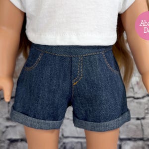 May include: Dark blue denim doll shorts with rolled cuffs and yellow stitching. The shorts have two front pockets and a waistband. The doll is wearing a white t-shirt. The shorts are designed for a doll.
