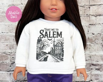 May include: A white long-sleeve sweatshirt featuring a black graphic print of a street scene with houses, trees, bats, and a cat. The text "Take me to SALEM" is printed in black above the illustration. The sweatshirt is worn by a doll with long dark hair.