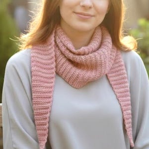 May include: A knitted pink scarf with a triangular shape and long, pointed ends. The scarf is made of a soft, textured yarn and is draped around the neck. The scarf is a light pink color.
